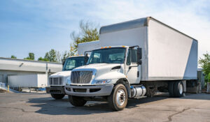 Two white box trucks parked side by side in a commercial lot near a warehouse building.